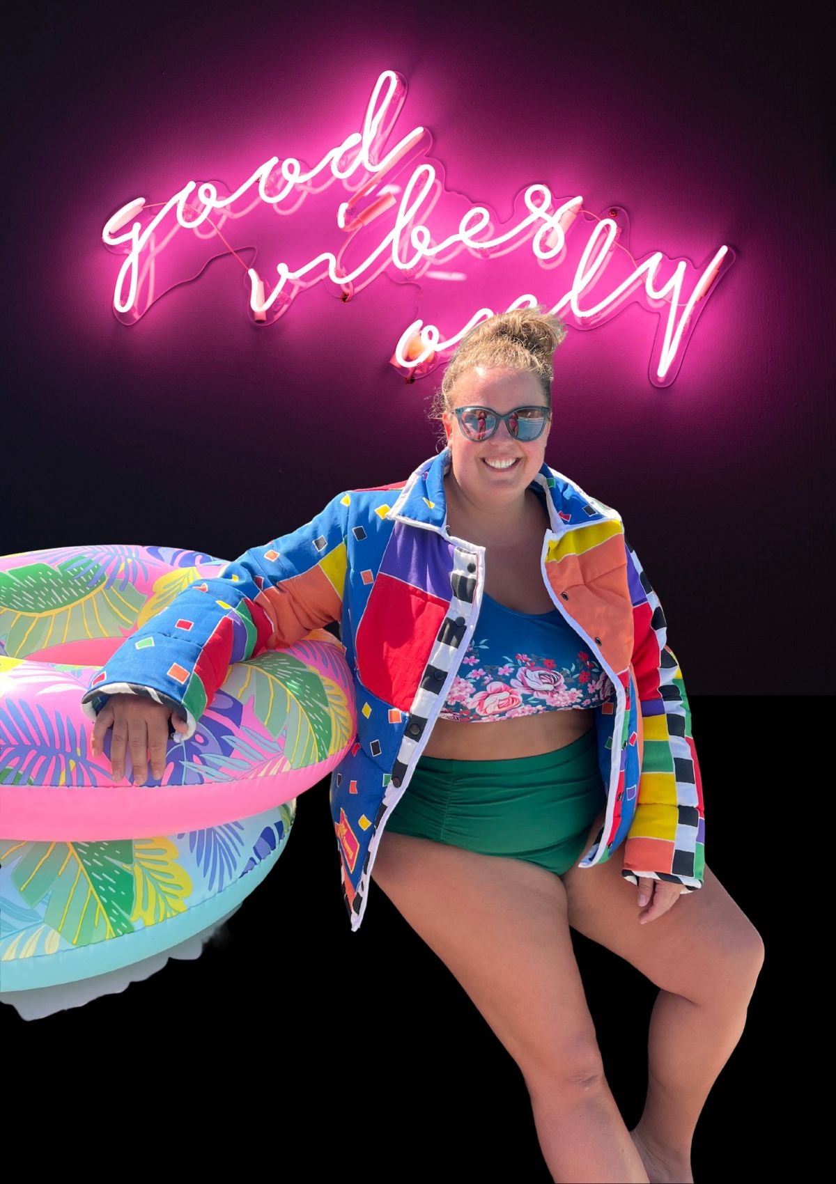 Person in colorful jacket upcycled from a comforter wearing green and blue bathing suit holding a pink inflatable ring with 'good vibes only' neon sign in background.