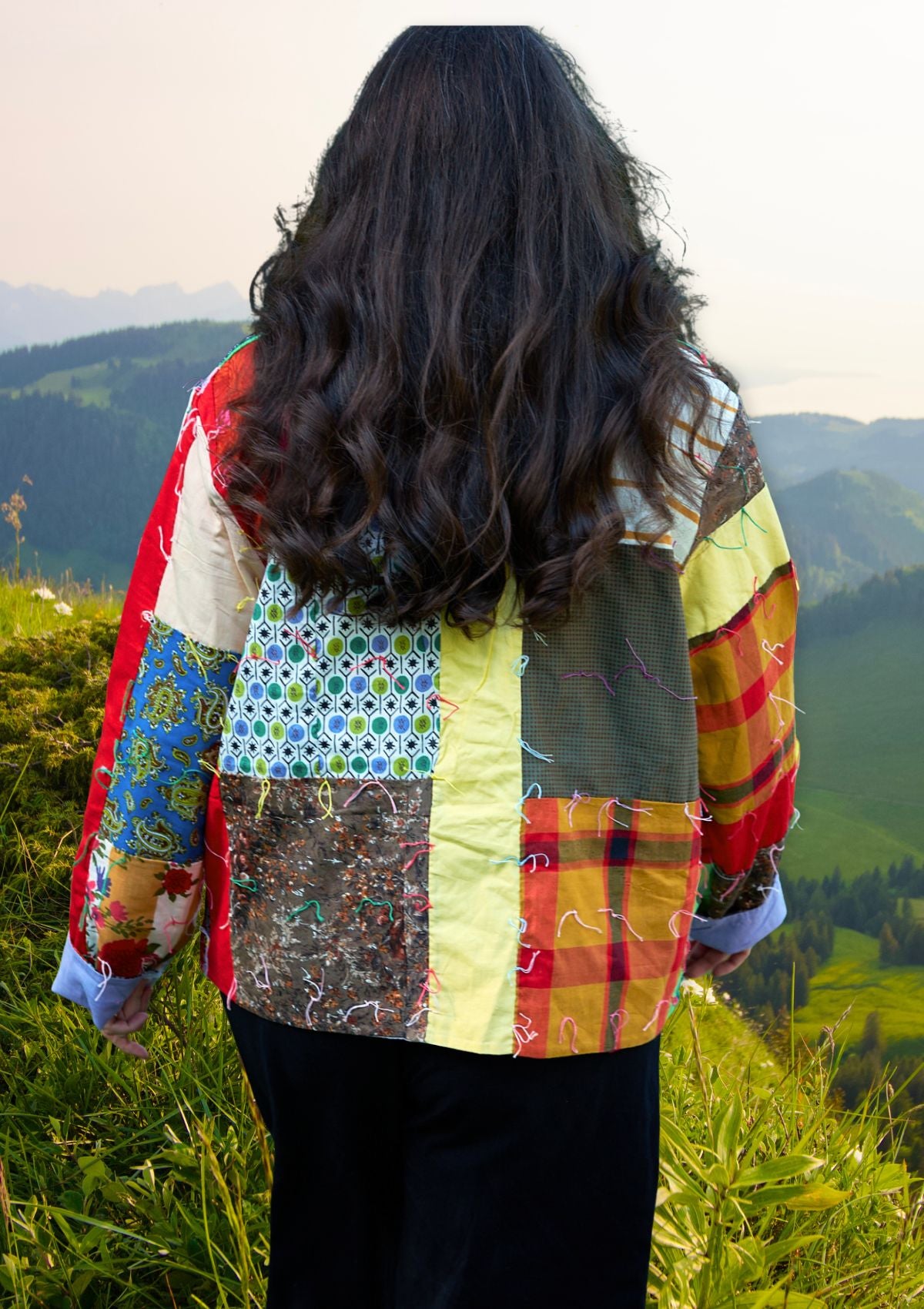 Person wearing a colorful patchwork jacket upcycled from a quilt standing in a scenic outdoor setting with mountains.