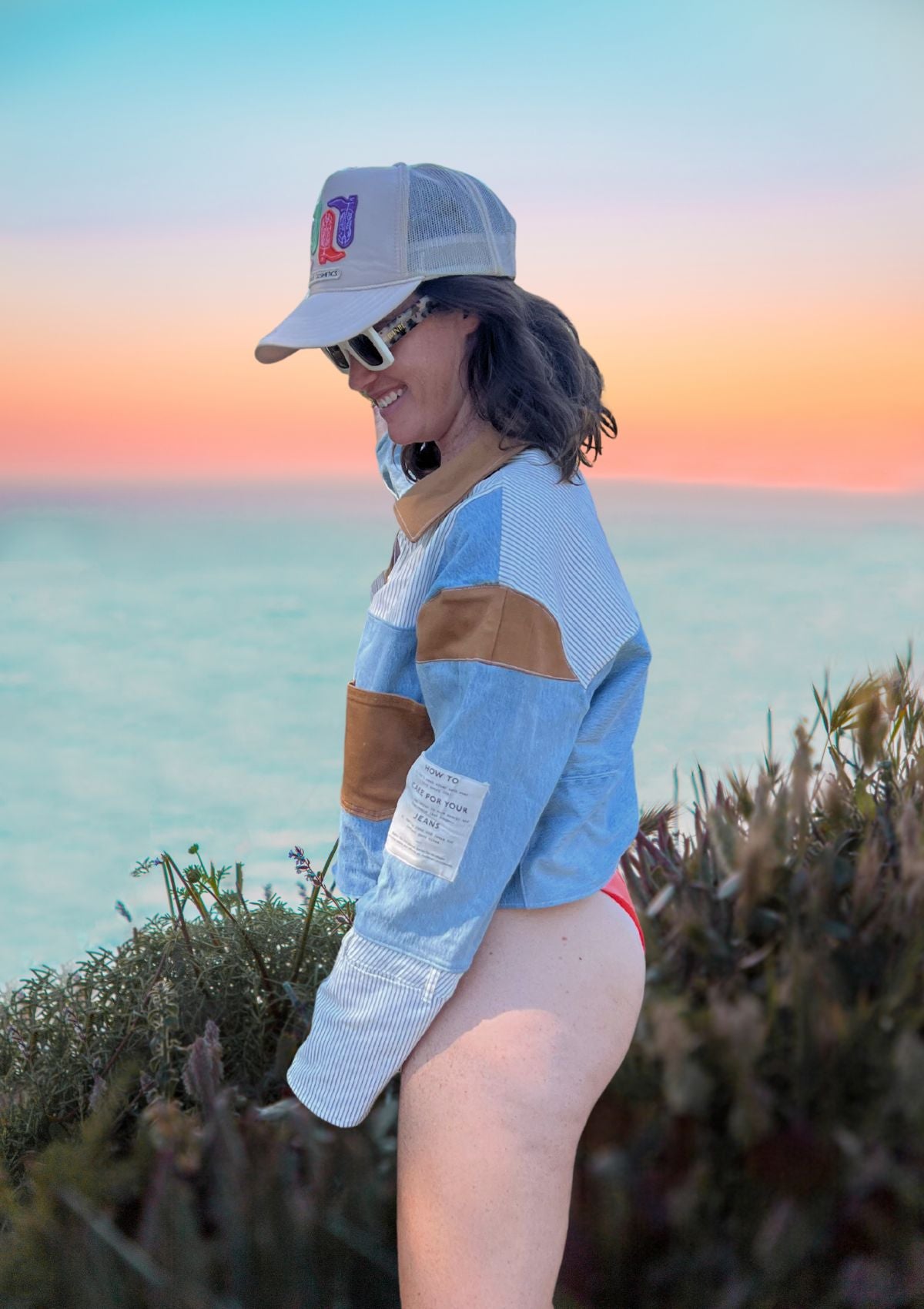Person wearing a denim patchwork jacket, bathing suit and cap with a sunset and ocean background.