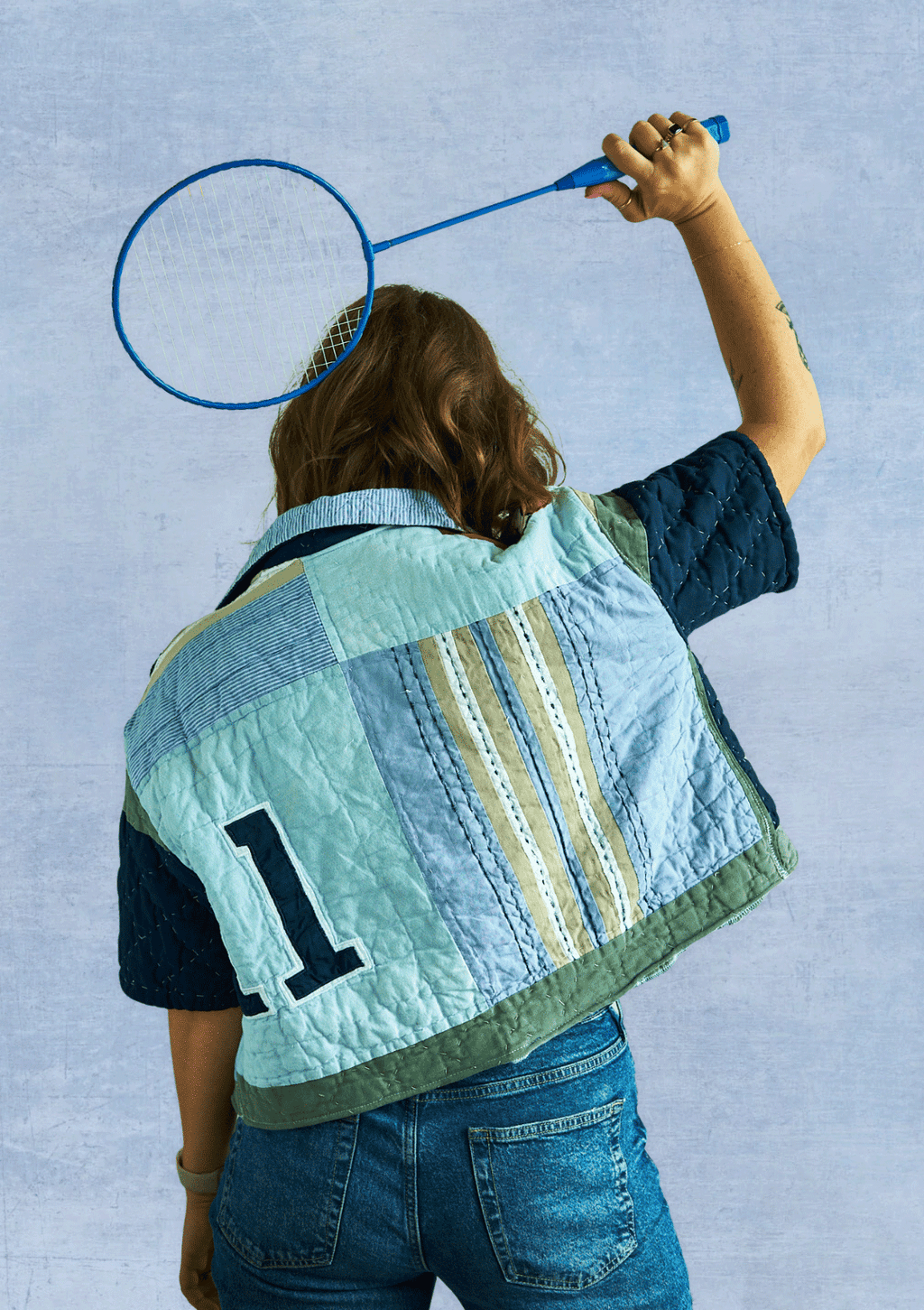 Person wearing a colorful quilted jacket, upcycled from a quilt with a badminton racket on a light blue background